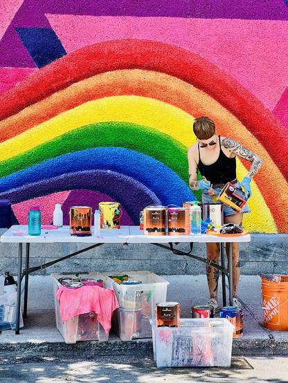 The image depicts a person working on a colorful mural featuring a rainbow. They are standing at a table filled with various paint cans and supplies, wearing gloves and focused on their task. The vibrant background adds to the artistic atmosphere of the scene.