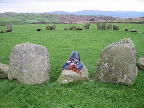 A photo of a stone circle with hills in the distance. The grass is a lush green colour. On one of the stones that has fallen down, I am laying-a white woman with long read dreadlocks in a denim jacket and jeans, and a striped scarf. I am looking up at the sky.
