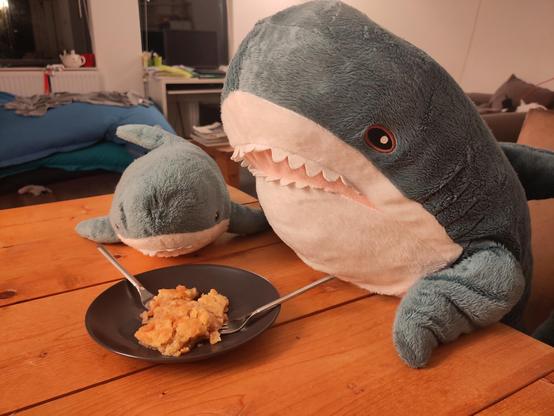 A Smolhaj cuddly shark and a Blåhaj cuddly shark sitting at the table. In between them is a plate with apple crumble and two forks.

They are lucky there is some apple crumble left, one of their humans ate almost all of it during a game night!