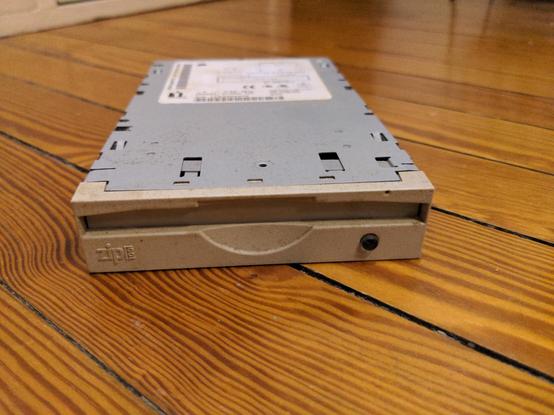 A 3.5" internal Zip drive lying on a wood floor. zip100 is molded into the plastic on the front and there's a translucent round eject button at the lower right. A white label is visible on the top, but out of focus.