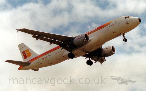 Low side view of a white, twin engined jet airliner flying from left to right at a very low altitude, with undercarriage lowered and flaps extended from the rear of the wings, suggesting it is about to land.
There is a thick red and orange stripe running along the body from over the top of the forward fuselage  to the tailcone at the rear.
There are white "Iberia" titles on the upper fuselage, repeated in red on the engine pods.
The tail has a stylised red lower-case letter "I" and a yellow upper-case letter "B", with a yellow crown in the dot over the "I".
The registration "EC-FMN" is on the top of the tail and the lower rear fuselage in black.
Cloudy sky with patches of blue fill the rest of the frame.