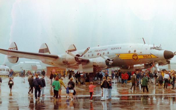 Side view of a 4 propellor-engined, triple-tailed, military transport aircraft in a bare metal finish, parked facing to the right.
There is a yellow flash on the nose with a black wireframe globe and black "MATS"  titles under the nose, and black "Military Air Transport Service" titles on the upper fuselage.
The tips of the propellors have red, white, and blue stripes, which will make concentric coloured circles when the engines are running.
Crowds of people are gathered around and under the plane, some walking around, others standing and looking.
The concrete apron they are on is saturated with rain water, puddles reflecting the people and plane.
Grey sky fills the top of the frame.