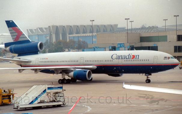 Side view of a grey, 3 engined jet airliner with a dark blue belly and engine pods, and a grey and red stripe running along the body, taxiing from left to right in front of a grey terminal building.
The plane has dark blue "Canadian" titles on the upper forward fuselage, the last letter "a" replaces with a red arrowhead overlaid on 5 grey horizontal bars.
That same logo appears in a larger form on the dark blue tail.
A white set of mobile airstairs is parked on the left of the frame in the foreground, with the wing of another plane on the right.
A grey terminal building stretches across the background, with a large grey hanger with pale blue doors is visible on the left.
