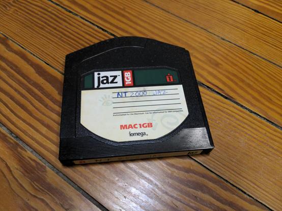 An Iomega Jaz disk lying on a wooden floor. The label reads MAC1GB with a handwritten NT 2000 JAZ in blue ballpoint pen.