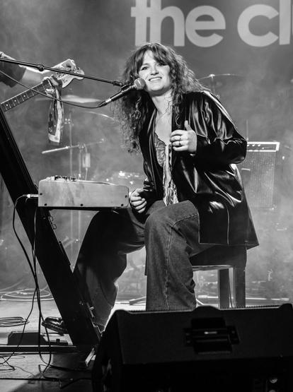 Black and white photo of a young woman sat on stage at a keyboard, smiling nd talking to the audience