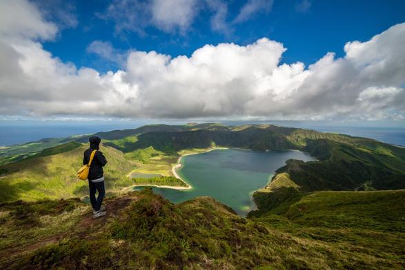 The image shows a stunning, expansive landscape. In the foreground, a person wearing a black jacket with a hood and carrying a yellow shoulder bag stands facing a large, green volcanic caldera with a turquoise lake at its center. The lake is surrounded by lush green hills and gentle mountain slopes covered with grass and scattered shrubs. In the distance, the blue sky is partially covered by dramatic, large clouds, casting light and shadow across the scenery. The image conveys a sense of tranquility and vastness, showcasing untouched nature.