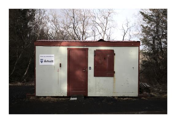A small, weathered worksite container with a red metal door and a rusted metal shutter on the front, surrounded by bare trees. A sign on the left side reads "Árholt" with a logo and Icelandic text. The setting is outdoors, with dark gravel and a wooden pallet on the right.