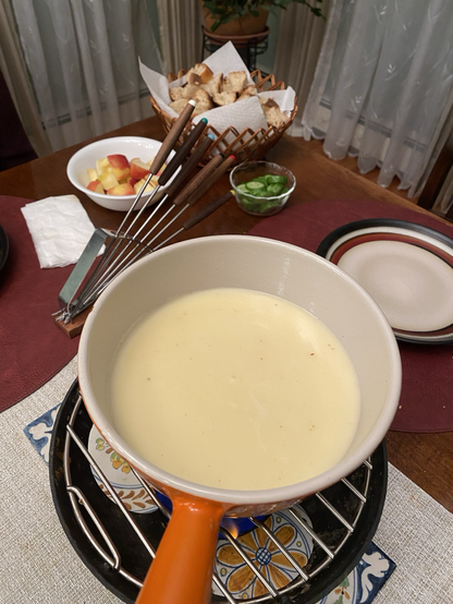 Photo shows a bit of bubbling hot cheese and wine and kirschwasser  a bowl if tii pi dated bread and apple in the background. A selection of long forks with which to dip said bread into said cheese