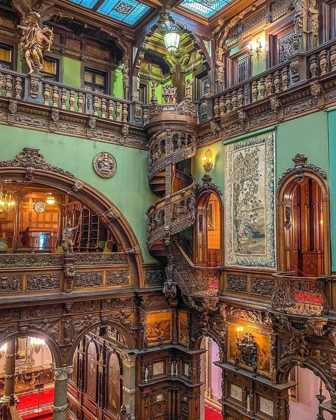 An ornate interior view of a grand hall, featuring intricate architectural details and rich wood carvings. Dominating the scene is a stunning spiral staircase with intricate wooden balustrades, rising elegantly towards the upper levels. The hall is adorned with detailed wooden arches, sculpted columns, and vibrant green walls. Stained glass panels in the ceiling allow soft, colorful light to filter in, illuminating elaborate wall tapestries and statues. The craftsmanship throughout reflects a blend of Gothic and Renaissance influences, evoking a lavish, historic ambiance.