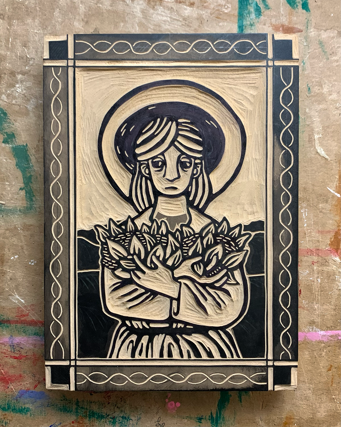 Photo of a carved linoleum block. Design is a frontal view of a medieval farmer holding plants in his arms. The farmer has a halo and is surrounded by a patterned border.