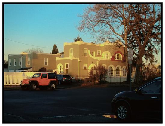 A grainy, colorful photo of a small apartment building (sorry i dont know architecture). an orange jeep rests outside towards the left of the image, and on the right a car can be seen close to the viewer, about to turn into the street. There is a tree that slightly frames the apartment building, and a bright early morning sun casts a golden glow across the building and the orange car. the trees almost look red as a result.