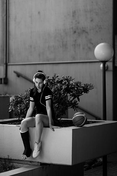 girl listening to music via headphones sitting on a small wall outside, with a basketball next to her. she wears a fiber walking cast on her right leg