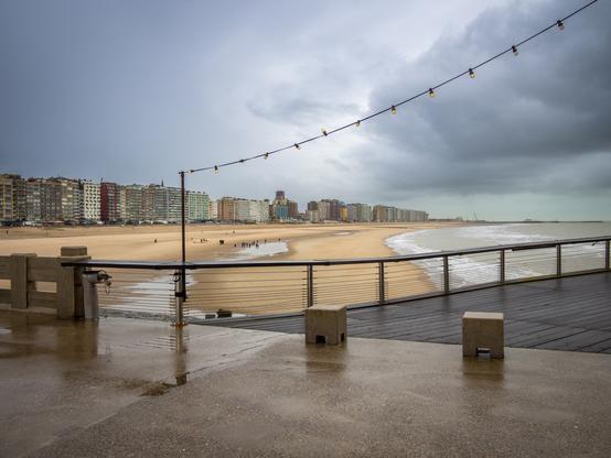 The image shows a beach scene under a cloudy sky. In the background are a row of buildings along the beach. The beach is empty, and the sea is visible on the right side of the image. A pier runs along the right side of the image. A string of lights hangs above the pier. The pier itself is wet from the rain. The foreground is a wet, concrete surface.
