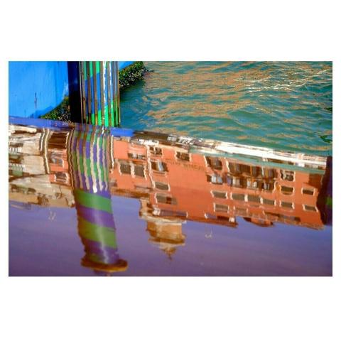 Reflet coloré d’un bâtiment vénitien dans l’eau, créant une image floue et vibrante. Les nuances d’ocre et de rouge du bâtiment contrastent avec les teintes bleues et vertes des poteaux et de la surface ondulante de l’eau. Ce jeu de reflets déforme les lignes architecturales et évoque une scène picturale presque abstraite.