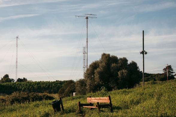 View of a empty bench, with a bin next to it. Behind them three huge antennas.
