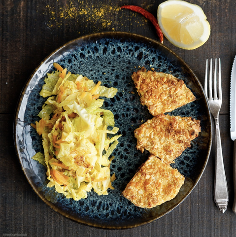 Dunkler Holzuntergrund. Runder, blau gesprenkelter Teller. Auf dem Teller: Spicy geröstete Sellerie-Nuggets in einer Panade aus Salzcrackern und Cheddar mit asiatischem Wirsing.
