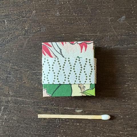 A miniature book with a cover made using colorful chiyogami paper is shown on a wooden surface, accompanied by a matchstick for scale. A band made from white washi paper with a dotted pattern surrounds the book.