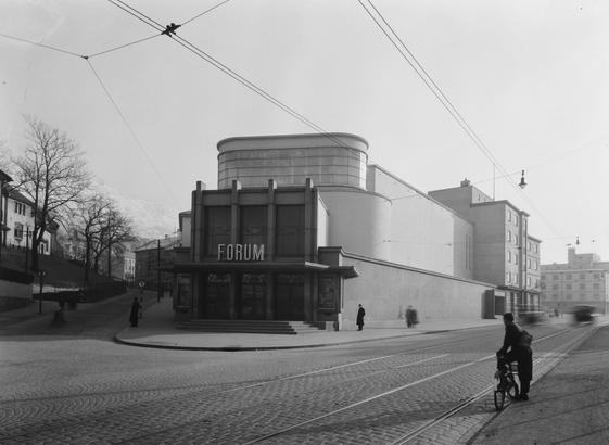 A black and white photo of a majestic art deco building with clean lines, leading down to a entrance where the marquee reads "FORUM". A man on a bike going past.