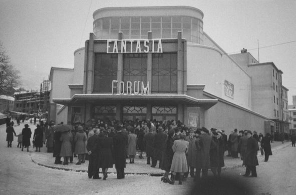 A black and white photo of a majestic art deco building with clean lines, where the marquee reads "FANTASIA" "FORUM". A crowd gathered around the snow on the street around the entrance, in warm clothing.