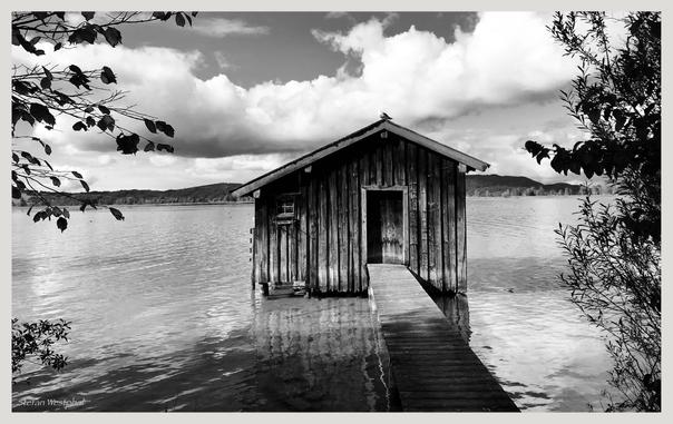 Eine Holzhütte steht auf Stelzen über ruhigem Wasser, verbunden durch einen langen Holzsteg. Die Szene ist in Schwarz-Weiß gehalten und zeigt einen wolkenverhangenen Himmel und entfernte Hügel. Das Laub in der Umgebung verleiht dem Bild einen natürlichen Rahmen.

A wooden cabin sits on stilts over calm water, connected by a long wooden walkway. The scene is in black and white, featuring a cloudy sky and distant hills. Surrounding foliage adds a natural frame to the image.