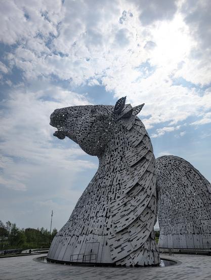 A monumental metal sculpture of a horse head raised to the sky, where the sun shines through the clouds. Part of the second horse head is visible behind it.