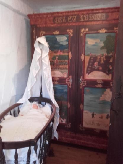An oval wooden baby cradle with white covering and a doll in front of a colourfully painted cupboard. The cupboard shows scenes of country life during the four seasons. The photo is poorly exposed and blurred because the room is darkened. We can't wake up the doll and the tired museum ghosts!