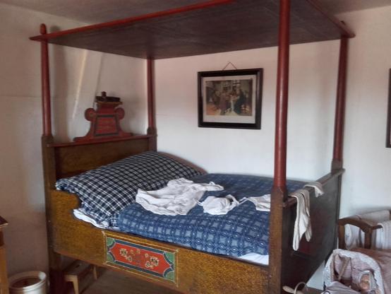 An old Alsatian four-poster bed with wooden canopy and carving with a shelf for a book at the headboard. The wood structure is an imitation potato print. This was used to make cheap wood appear valuable. Colourful peasant paintings decorate the side. The bedding is covered with traditional blue kelsch, a chequered linen that used to be dyed with plants from the garden. The canopy was made of wood because people slept in unheated rooms. Therefore, pieces of plaster often fell from the ceiling. The wood protected the sleepers and curtains kept the warmth of the hot water bottles in bed for longer.
The chamber pot beside or under the bed was a help, too. These families had only one extern outhouse (pit latrine) in the yard. Imagine the long walk outside in winter!