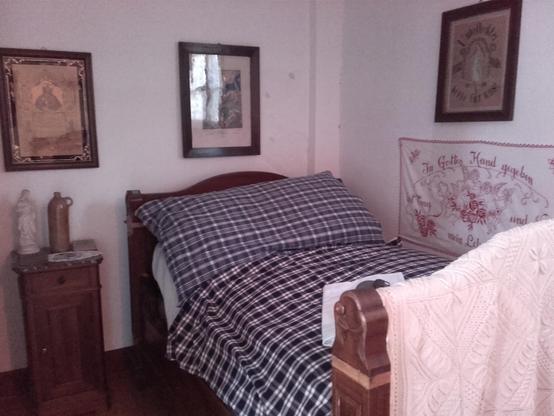 Simple Alsatian bed with blue chequered Kelsch cover in an alcove, surrounded by religious pictures. The statue of the Virgin Mary on the bedside table was as important as the hot water bottle, which was often made from empty shell casings during the First World War.
The hand-embroidered wall hanging was not only a religious and folk-magical protection, it also insulated the cold walls. Alcoves were extensions of the only heated parlour. The curtains remained open during the day. When people went to bed, they closed them.