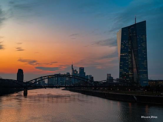 Eine malerische Abendaufnahme von Frankfurt am Main, die die imposante Skyline der Stadt und den markanten EZB-Turm in sanftes Licht taucht. Der orangefarbene Himmel spiegelt sich im ruhigen Wasser des Mains, während die Brücke elegant über den Fluss führt und die Lichter der Stadt in der Dämmerung leuchten.