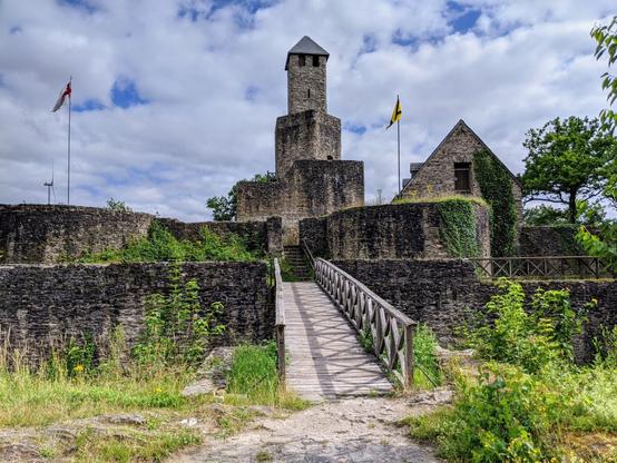 Stone castle ruins with a central tower and connecting walls, featuring a wooden bridge leading to an entrance. Flags fly atop the tower, and greenery grows on the stone structures under a cloudy sky.