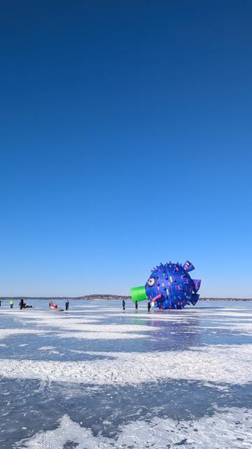 A scene on frozen Lake Mendota. The sky is completely clear and the ice is smooth, with some patches of snow. On the ice is a strangely shaped blue object, resembling a mix of a hot air balloon or spherical kite. Maybe it's meant to represent a bacterium or virus? Some people are standing on the ice next to it.