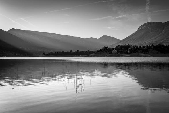Black and white photography i which you can see a lake in calm with some reflections of the mountains at the background. Also, in the mountains there are a smooth light of the late afternoon.