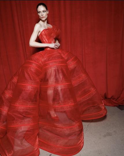 A model poses in a striking, voluminous red gown made of sheer, structured fabric. The strapless bodice is pleated with a sculptural ruffle on the chest, cinching at the waist before cascading into a dramatic, bell-shaped skirt. The transparency of the fabric reveals her legs, adding an element of modern sensuality. She wears red pointed-toe heels, blending seamlessly with the monochrome red background.