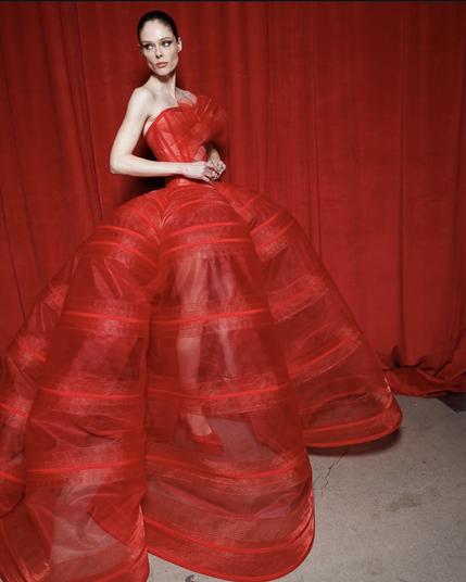 A model poses in a striking, voluminous red gown made of sheer, structured fabric. The strapless bodice is pleated with a sculptural ruffle on the chest, cinching at the waist before cascading into a dramatic, bell-shaped skirt. The transparency of the fabric reveals her legs, adding an element of modern sensuality. She wears red pointed-toe heels, blending seamlessly with the monochrome red background.
