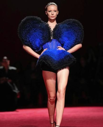 A model walks the runway in a bold black and blue mini dress with exaggerated, circular puffed shoulders resembling butterfly wings or ruffled clouds. The bodice is structured with a corseted silhouette, transitioning from an electric blue center to deep black edges. The short hemline highlights her legs, and she pairs the look with black heels. Her hair is slicked back, emphasizing the dramatic shape of the garment.