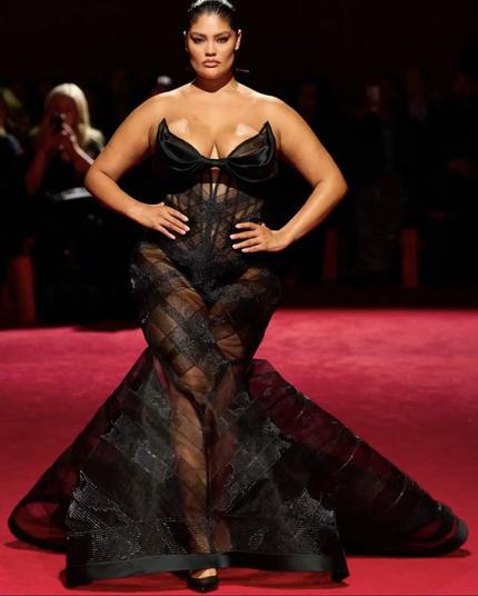 A curvy model confidently walks down the runway in a striking black mermaid gown. The dress features a sheer, crisscrossed panel skirt that flares out at the bottom, creating volume. The strapless bodice has a dramatic bow-like detail across the bust, adding a touch of femininity. The gown’s transparency reveals glimpses of her skin, blending sensuality with high-fashion drama.