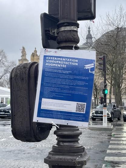 Une affiche imprimée en A4 et plastifiée, posée sur un feu de circulation ou on peut lire "Expérimentation vidéoprotection augmentée" puis un texte en tout petit