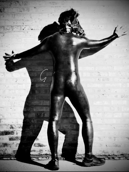 This black-and-white image captures a human pony clad in a sleek black silicone catsuit, emphasizing a second-skin aesthetic that enhances both anonymity and transformation. Their head is adorned with a leather horse hood, a key element of pony play gear that embodies the equine persona by mimicking the features of a horse, such as elongated snouts and decorative bridles.

The pose—hands splayed against a brick wall—evokes a sense of restrained power and submission, hallmarks of the dynamic between "pony" and "handler" in this kink. The interplay of light and shadow adds depth to the scene, highlighting the reflective texture of the catsuit while casting an equine-like silhouette that further immerses the viewer in the fantasy.

The leather hood and full-body suit serve as tools for escapism, allowing the individual to shed their human identity and embody something entirely different.