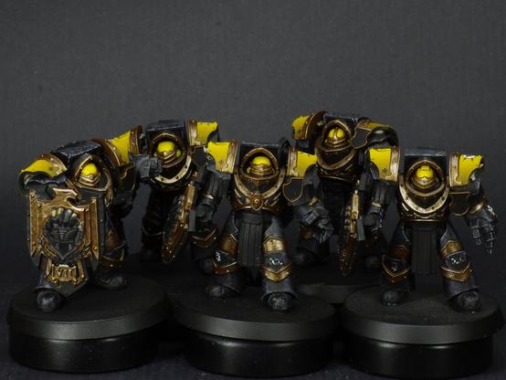 A squad of 5 Imperial Fist Huscarl terminators from the Horus Heresy. They're wearing black armour with yellow helmets and shoulder pads. They have heavy shields and power gauntlets.