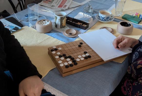 To people playing a 9x9 game of go on a kitchen table. The game board is self built and laserengraved. The stones and cups are 3D printed. Currently the board is folded in half and paper covers another quarter.