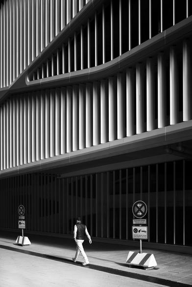 a pedestrian walks quickly in front of a parking garage with a striking façade (white vertical metal slats). with his light-colored jeans, he stands out very strikingly against the shadow of the parking garage.