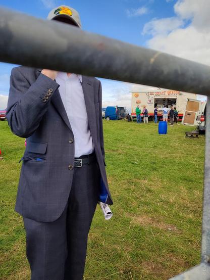 A medium shot of a person in a suit and cap standing in front of a snack wagon at an outdoor event, partially obscured by a metal barrier. The snack wagon says "THE SNACK WAGON" on top.