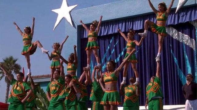 A cheerleading squad of Black teenagers in green cheer uniforms, forming a complex human pyramid on a stage.