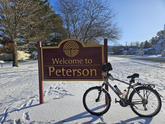 A bicycle leaning against a sign that says "Welcome to Peterson". The ground is covered in white snow, tire tracks and footprints visible. Trees, buildings, a blue sky and a grey street are visible on the background.
