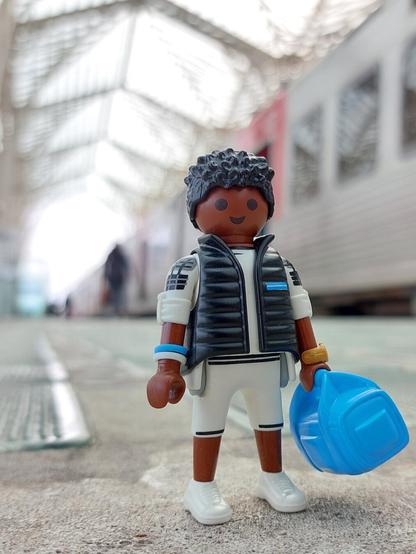 Jogador playmobil na estação de comboios gare do oriente em Lisboa. Cabelos aos caracóis pretos, casaco preto, calções brancos, tom de pele acastanhado, saco de mão azul, relógio dourado. Ao lado de um comboio urbano cinza com detalhes vermelhos