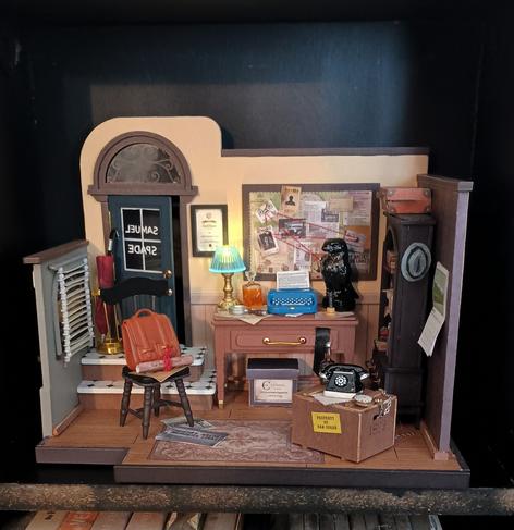 A picture of a model. It's a diorama of a room; specifically, a detective's office. A typewriter sits on a messy desk next to a recreation of the Maltese falcon statuette. There's a bulletin board covered in papers and red string hanging above the desk, and 'Samuel Spade' is painted in reverse on the glass pane of the door in the back of the room.