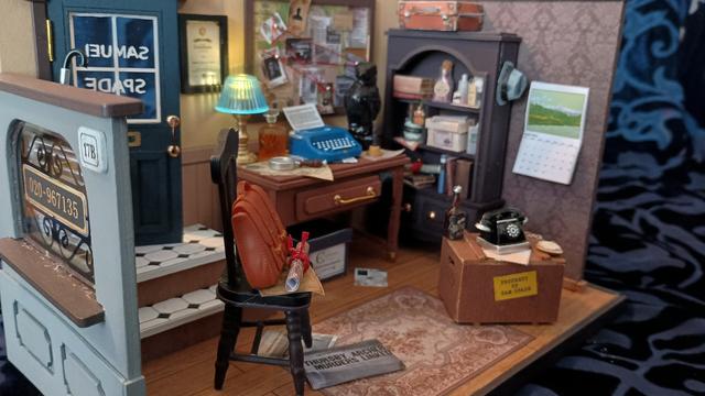 A picture of a model. It's a diorama of a room; specifically, a detective's office. The camera is focused on a chair. A leather satchel and some papers are placed on the chair. In the background are a desk; a tall bookcase; and a wooden crate that has 'Property of Sam Spade' printed on its side. All objects are covered in clutter and mess.