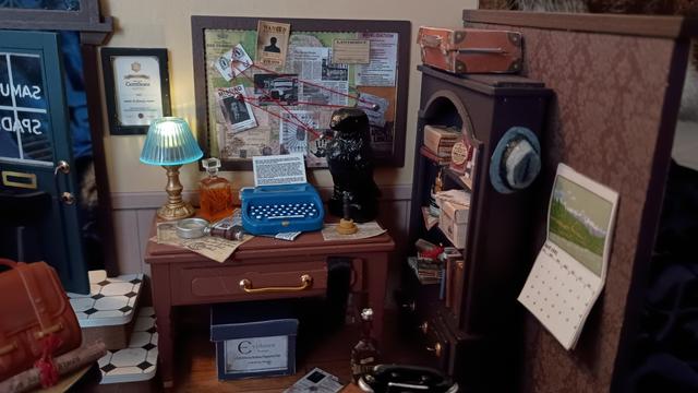 A picture of a model. It's a diorama of a room; specifically, a detective's office. A closeup on a messy desk covered in detritus: a lit, working lamp; a decanter of liquor; a magnifying glass; a typewriter with a piece of paper loaded into it and in use; and a replica of the Maltese falcon statuette. On the wall above the desk hangs a bulletin board covered in papers and red string, and a framed certificate.