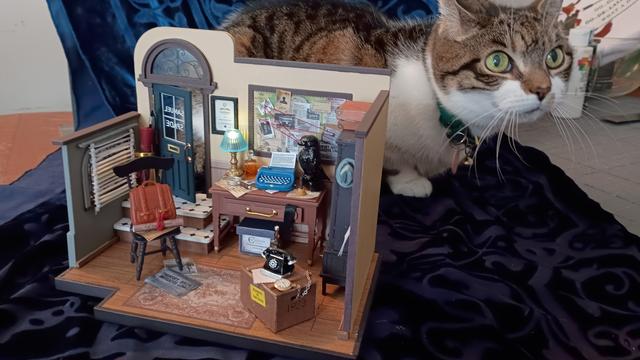 A picture of a model. It's a diorama of a room; specifically, a detective's office. A cat perches behind the model, dwarfing it in size.