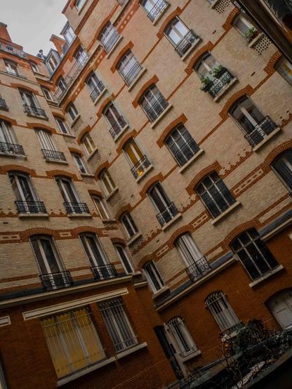 La photographie montre la façade d'un bâtiment résidentiel, caractérisé par un travail de briques avec un mélange de briques claires et rouges. Le bâtiment présente de nombreuses fenêtres avec des garde-corps décoratifs en fer forgé, typiques de l'architecture européenne plus ancienne. Le bâtiment est pris en contre-plongée depuis une cour intérieure.